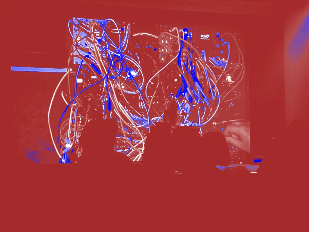 Keith Fullerton Whitman stands playing modular synth in front a projection of his rig